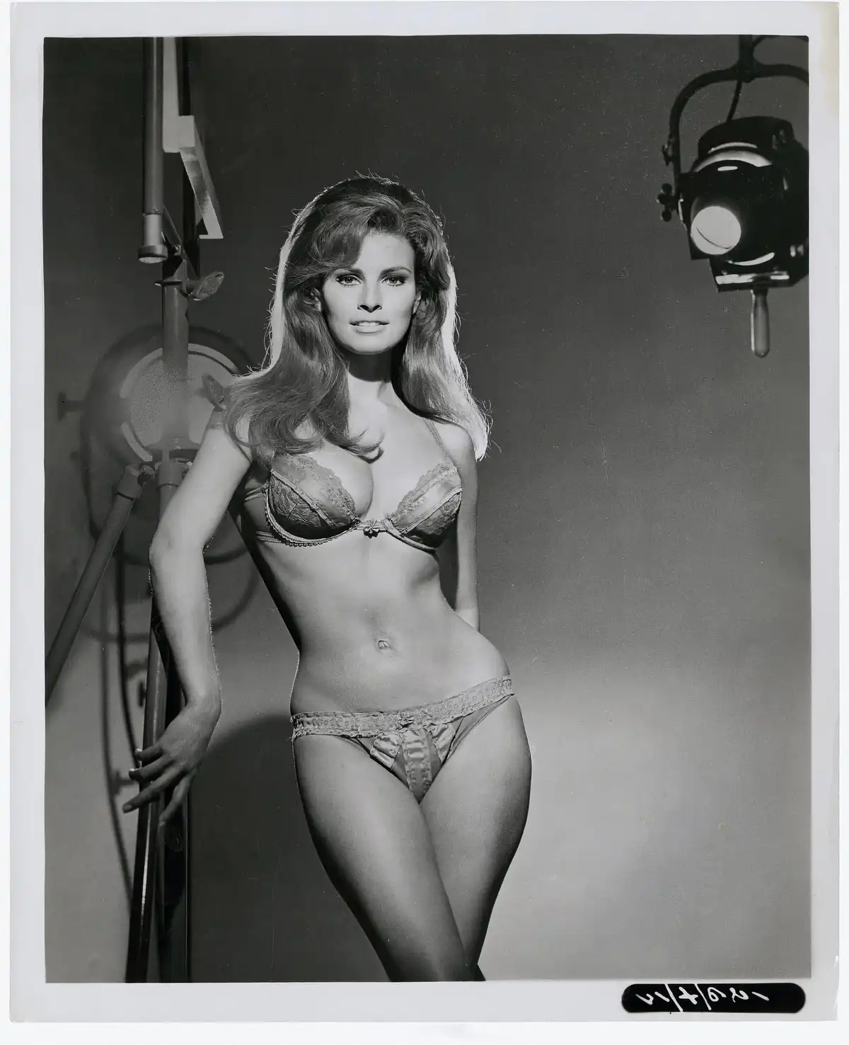 A black-and-white studio photograph shows a woman with long, styled hair posing in lingerie, standing next to a lighting rig…
