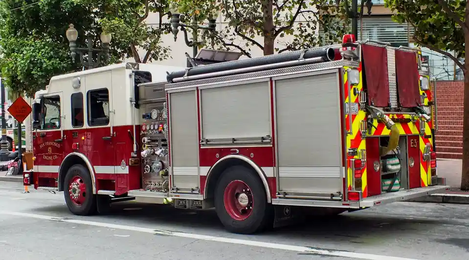 A red and white San Francisco fire engine is parked on a city street next to a tree, with a visible 'LEFT LANE CLOSED AHEAD'…