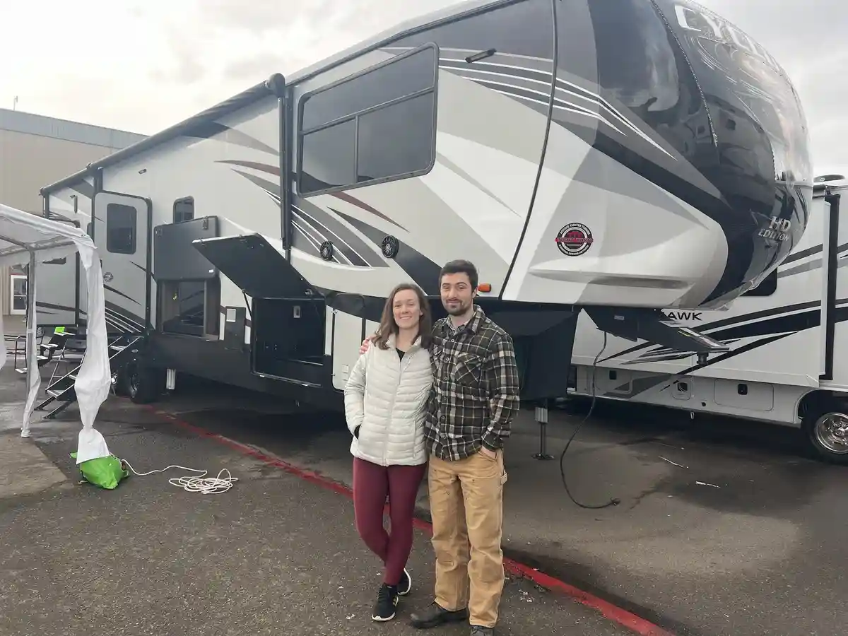 A man and a woman stand smiling in front of a large, modern RV with black, white, and gray graphics. The RV has the word…