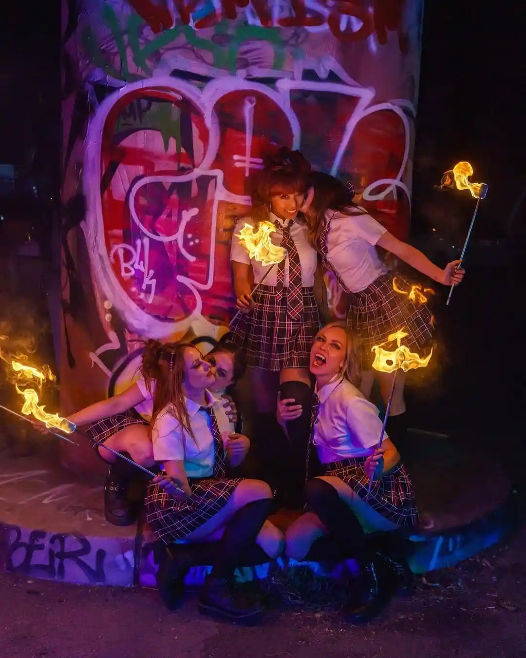 Four young women in school uniforms pose playfully around a graffiti-covered pillar at night, each holding a flaming torch…