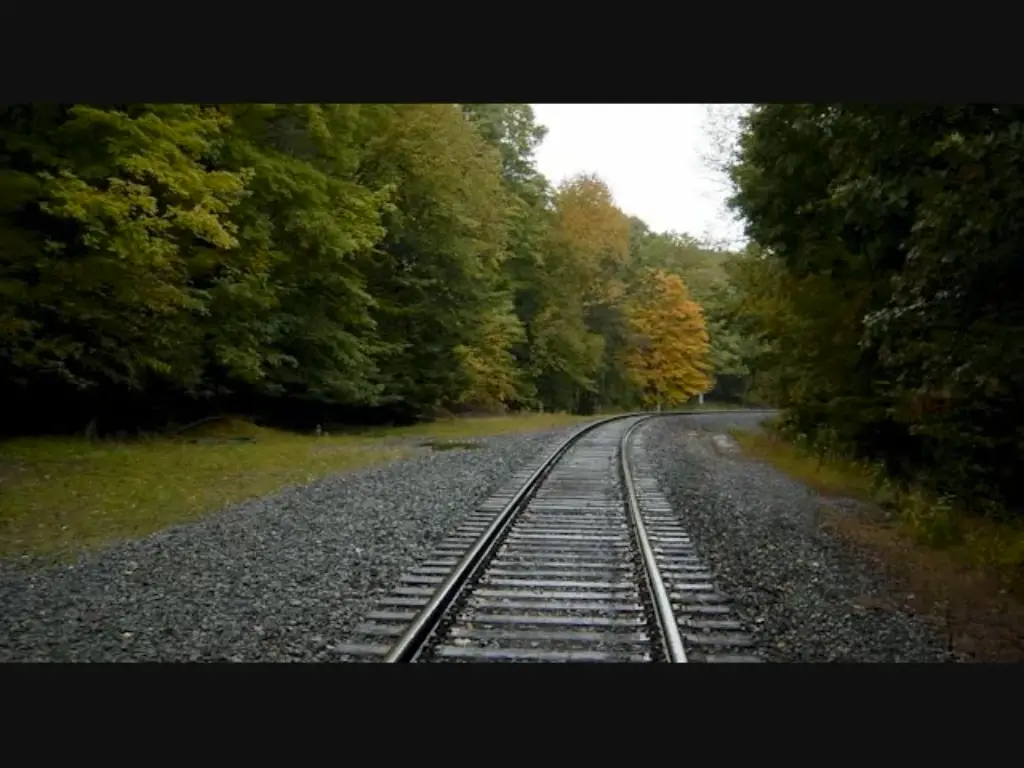 A photograph of a railroad track curving gently to the right through a forest with autumn-colored trees. The overlaid text…