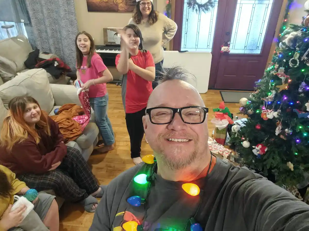 A man with glasses takes a selfie in a living room decorated for Christmas, wearing a string of colorful, lit-up holiday…