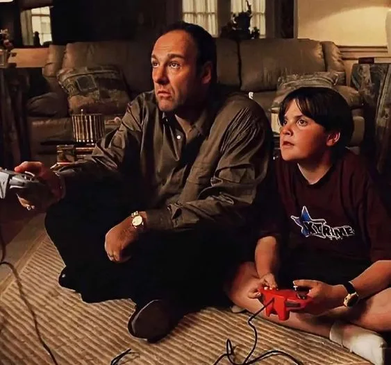 A man and a young boy sit cross-legged on a rug in a living room, both holding video game controllers and looking upward…