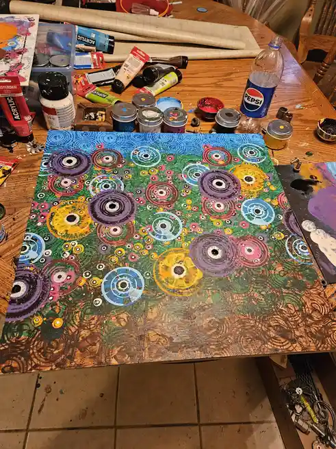 A colorful abstract painting with concentric circles in various hues sits on a cluttered wooden table surrounded by paint…