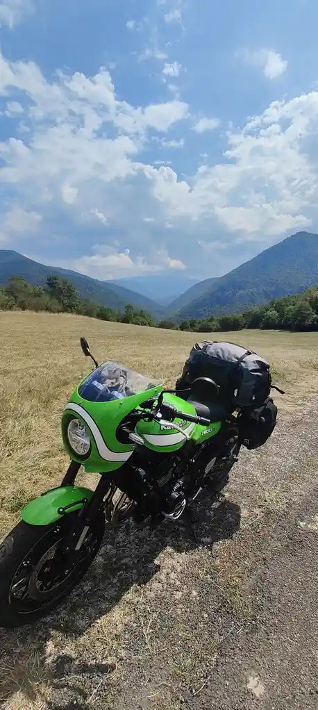 A bright green Kawasaki motorcycle with luggage is parked on a gravel path in a grassy field, with mountains and a partly…