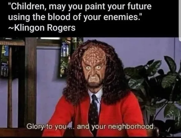 A man in a red sweater and tie wears a grotesque, textured mask with wild hair and an angry expression. The image has text…