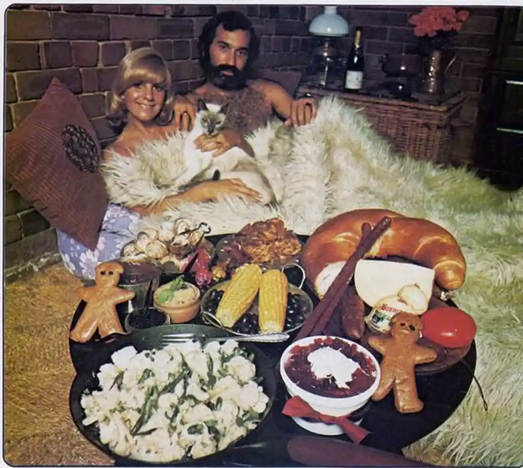 A vintage photo shows a man and woman lounging on a bed covered in a white fur blanket, holding a cat between them. In front…