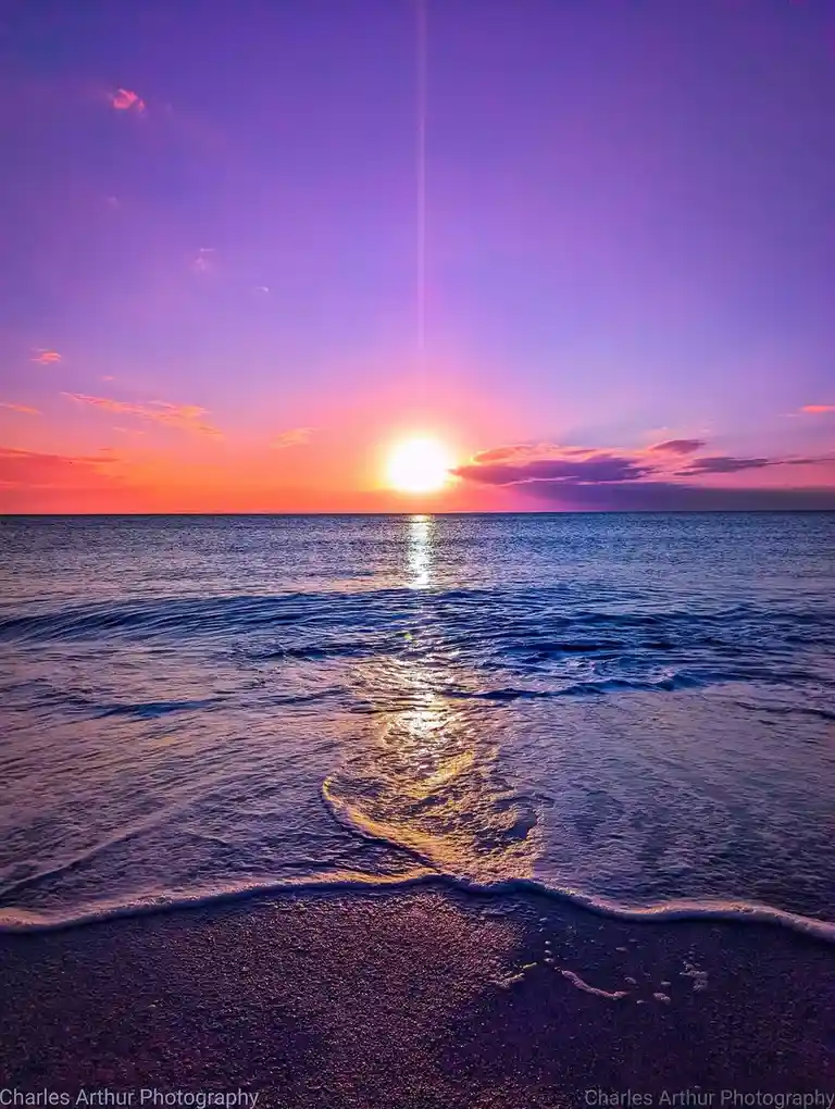 A vibrant sunset over the ocean with the sun low on the horizon, casting a purple and orange sky and a shimmering reflection…