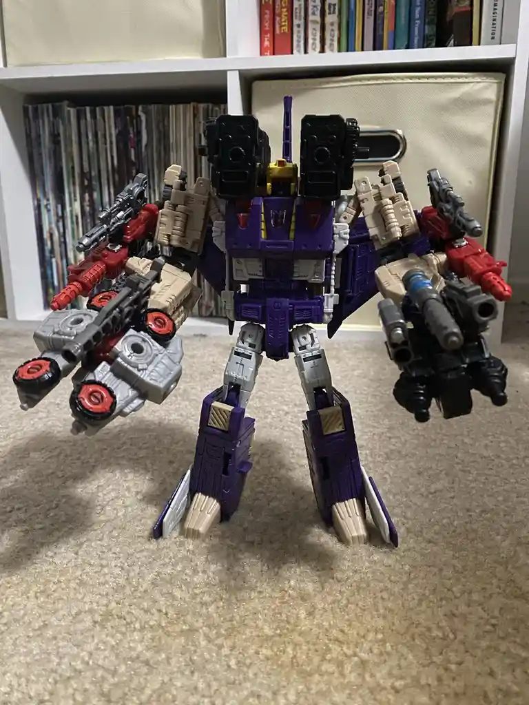 A large purple and beige Transformers toy robot stands on a beige carpet, heavily armed with multiple guns and cannons on…