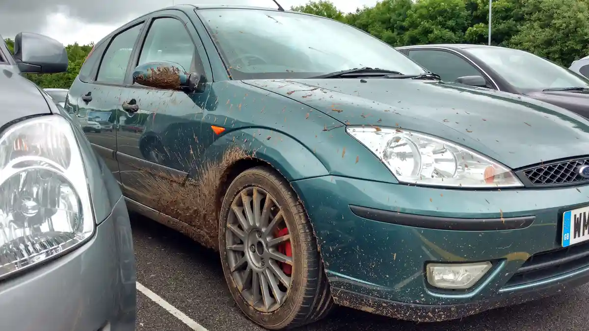 A teal Ford Focus hatchback is covered in dried mud splatters across its hood, doors, and side mirror, parked in a lot with…