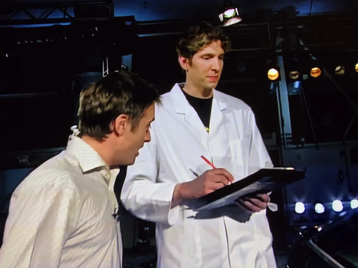 Two men are in a dimly lit stage or studio setting with visible lighting rigs. The man in the foreground, wearing a…