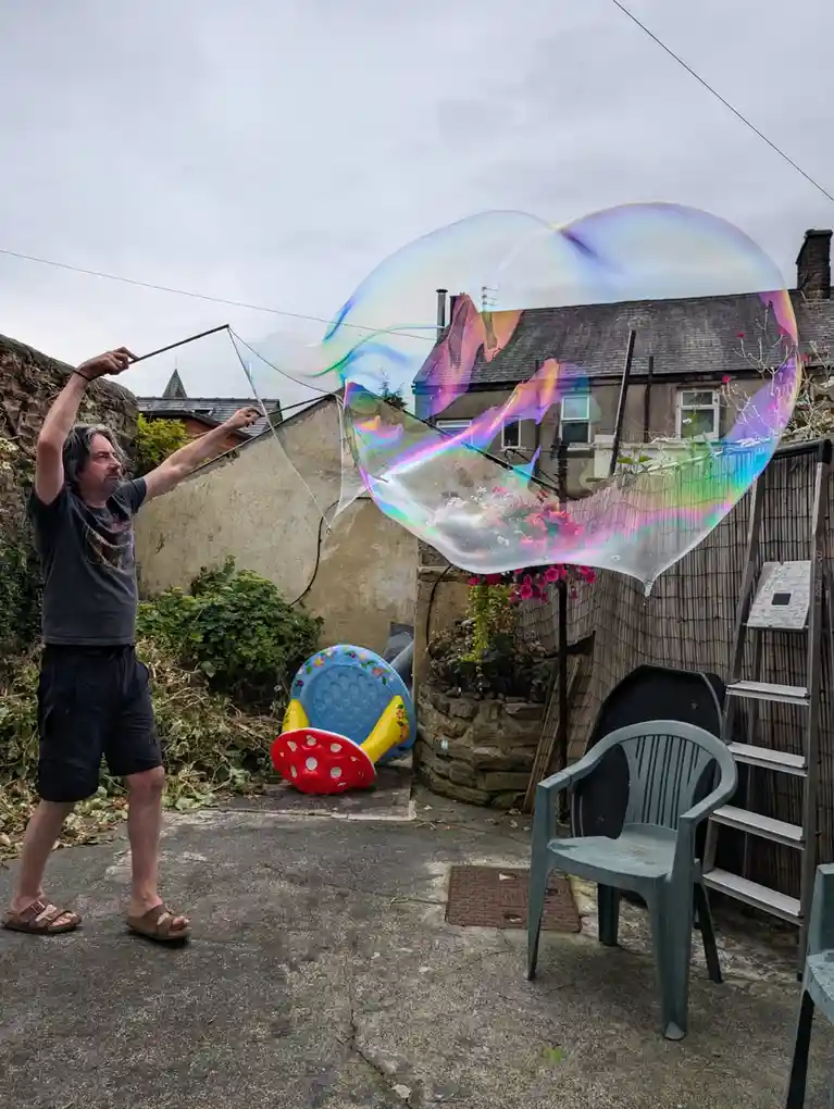 A man with gray hair and a beard stands in a backyard holding a bubble wand, creating a massive, iridescent soap bubble that…