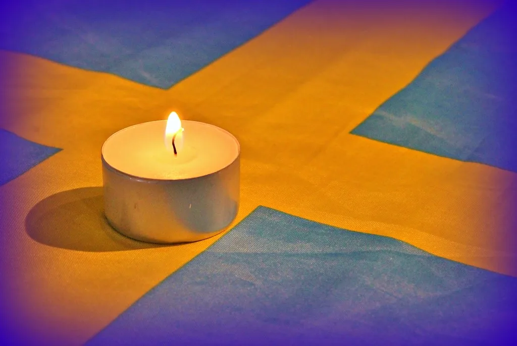 A single lit tealight candle sits centered on a Swedish flag, its warm glow illuminating the yellow and blue cross pattern…