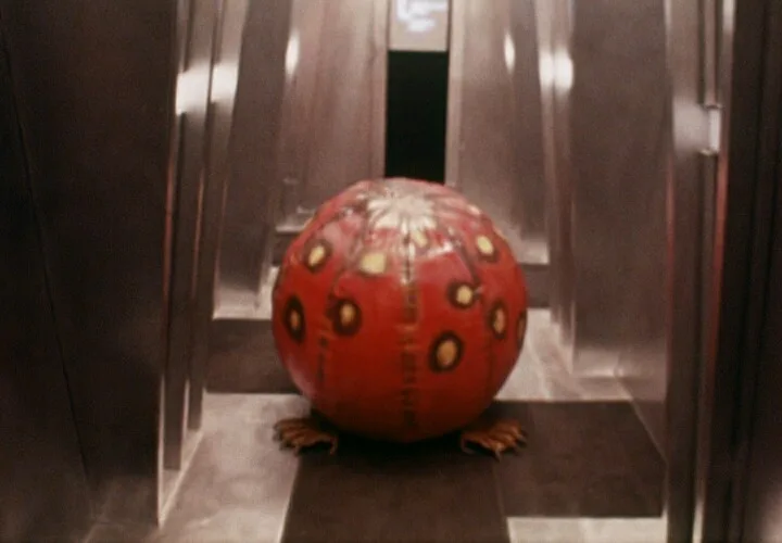 A red, spherical creature with yellow-ringed spots and small clawed feet sits inside an elevator with metallic walls. The…