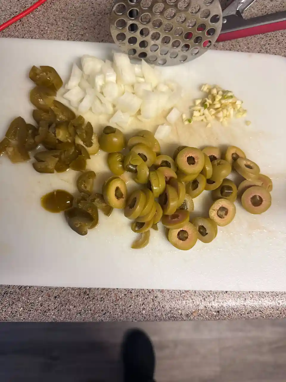 A white cutting board holds piles of chopped ingredients: diced white onions, minced garlic, sliced green olives, and…