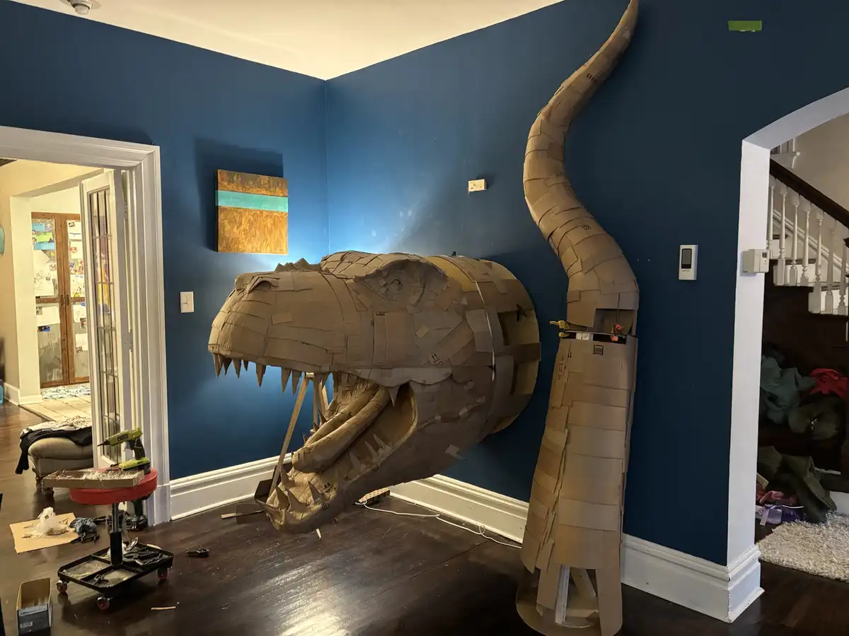 A large, life-sized T-Rex head and neck sculpture made entirely of cardboard stands in a blue-walled room, with tools and…