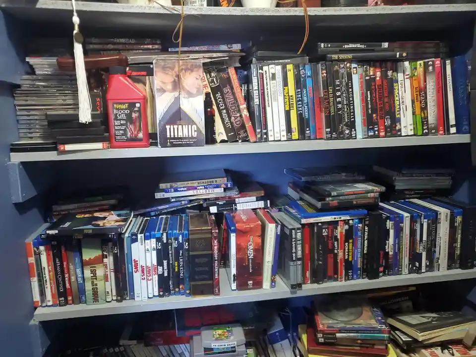 A cluttered shelf filled with DVDs and video games, including a red bottle of 'Spirit Blood Gel' next to a 'Titanic' DVD…