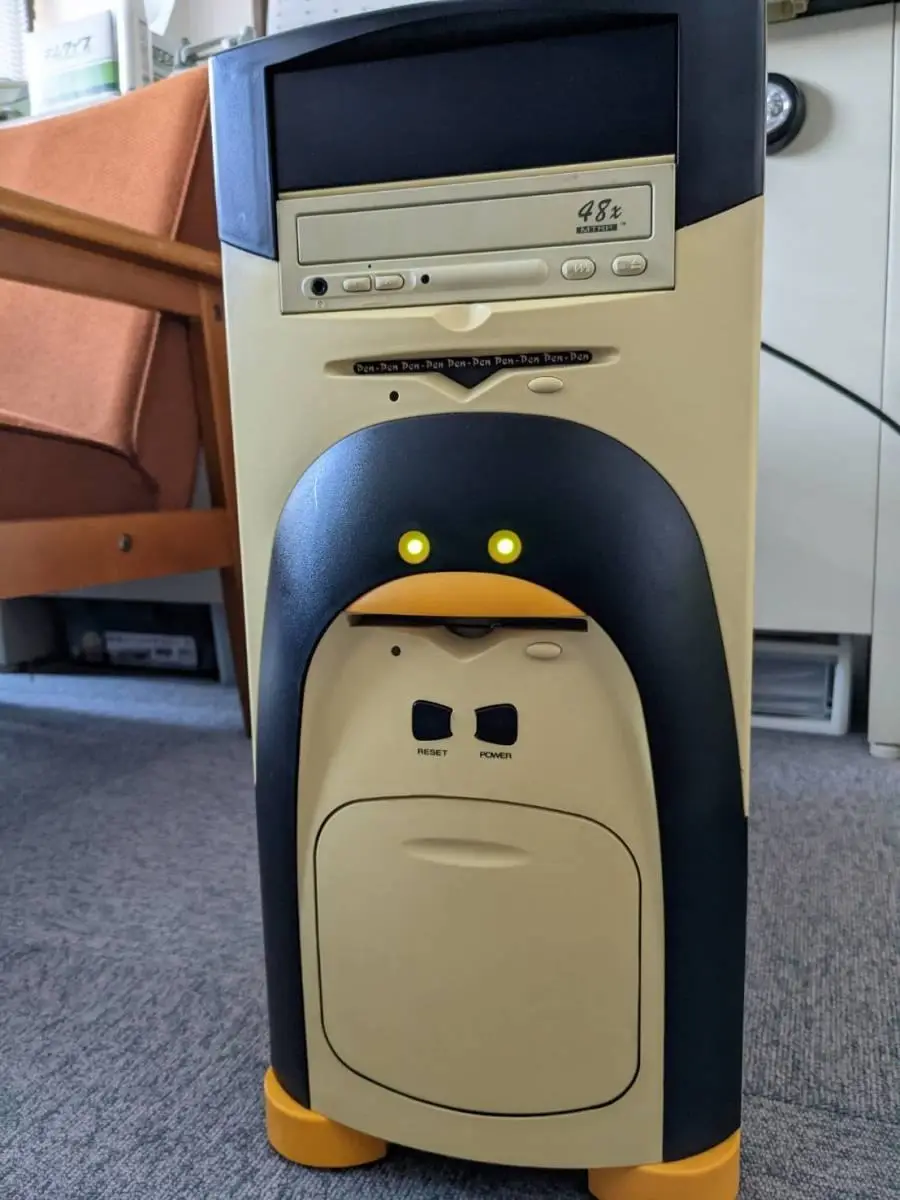 A beige and black desktop computer tower with a penguin face design, featuring glowing yellow eyes, an orange beak, and…