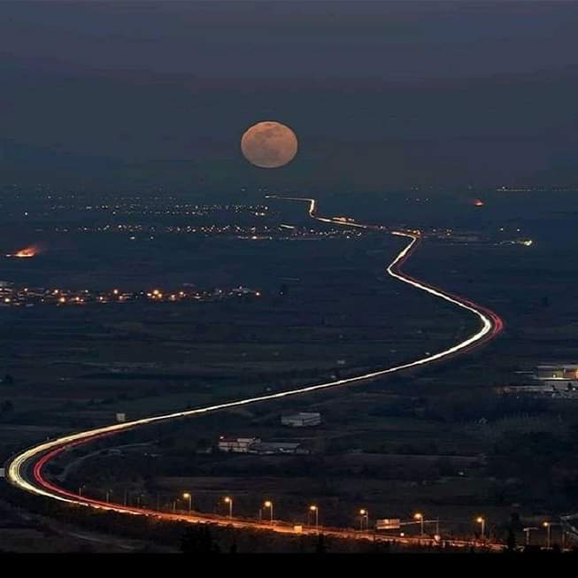A long-exposure night photo shows a winding road with light trails from cars, curving toward a large, full moon in the sky…