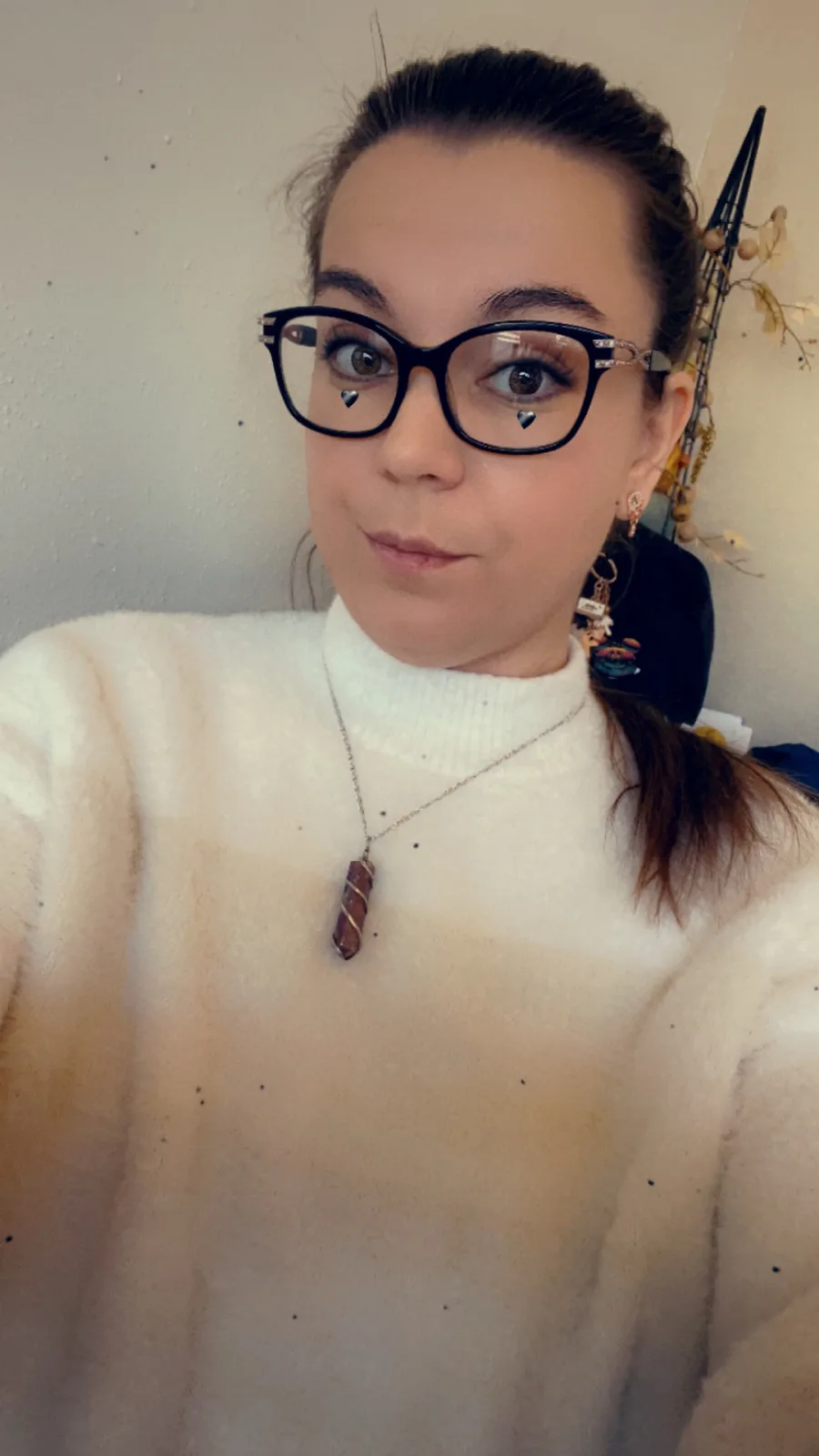 A selfie of a woman with dark hair pulled back, wearing black-rimmed glasses, a white fuzzy turtleneck, and a necklace with…