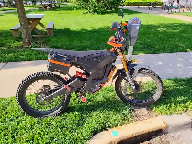An electric dirt bike with black and orange accents is parked on grass next to a sidewalk, with its front wheel wedged…