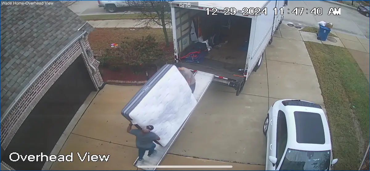 An overhead security camera view shows two men struggling to lift a large mattress up a ramp into the back of a white moving…