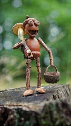 A whimsical, handcrafted figurine of a smiling acorn character with twig limbs, a leaf hat, and a small basket, standing on…