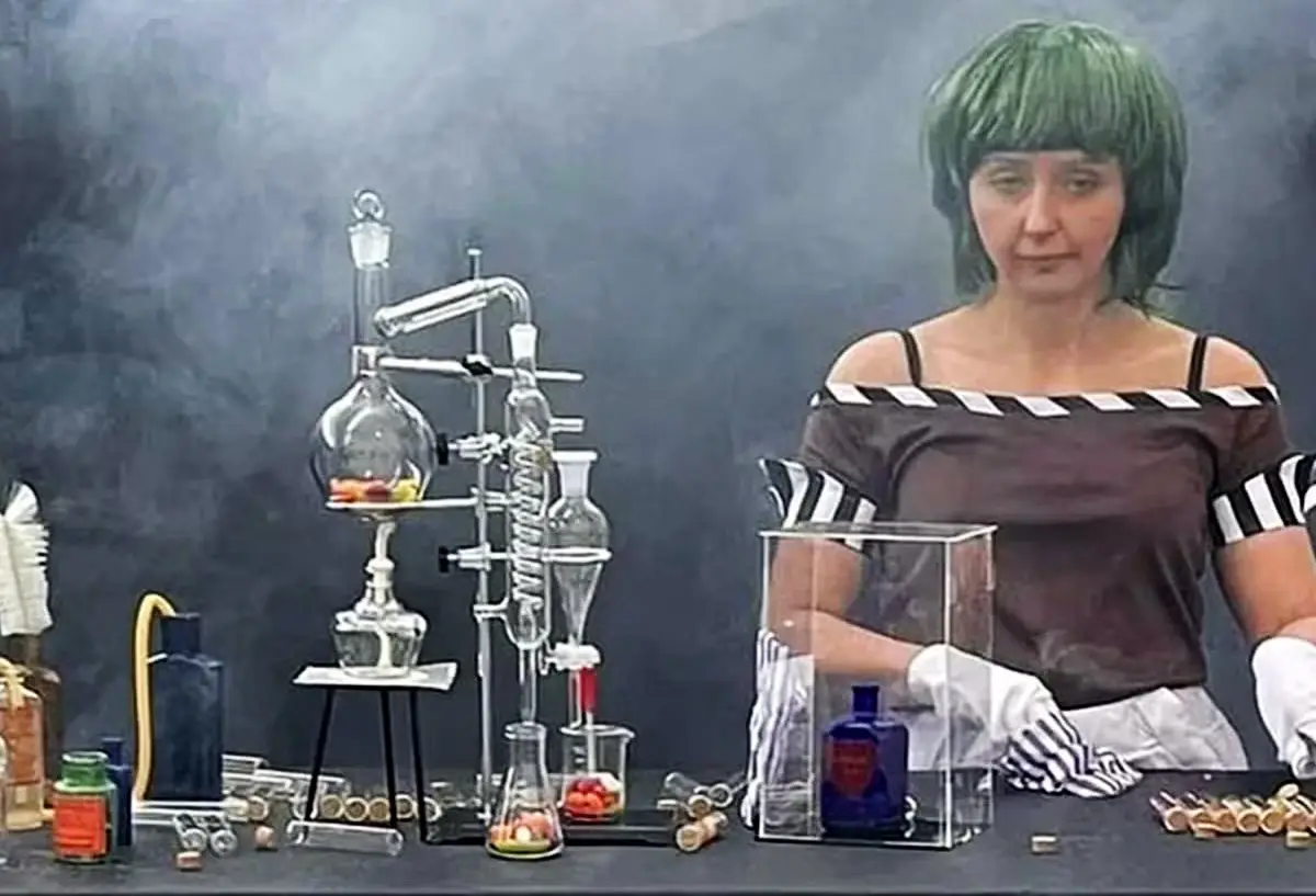 A woman with a green wig and striped shirt stands behind a lab bench filled with glassware, beakers, and smoking apparatus…