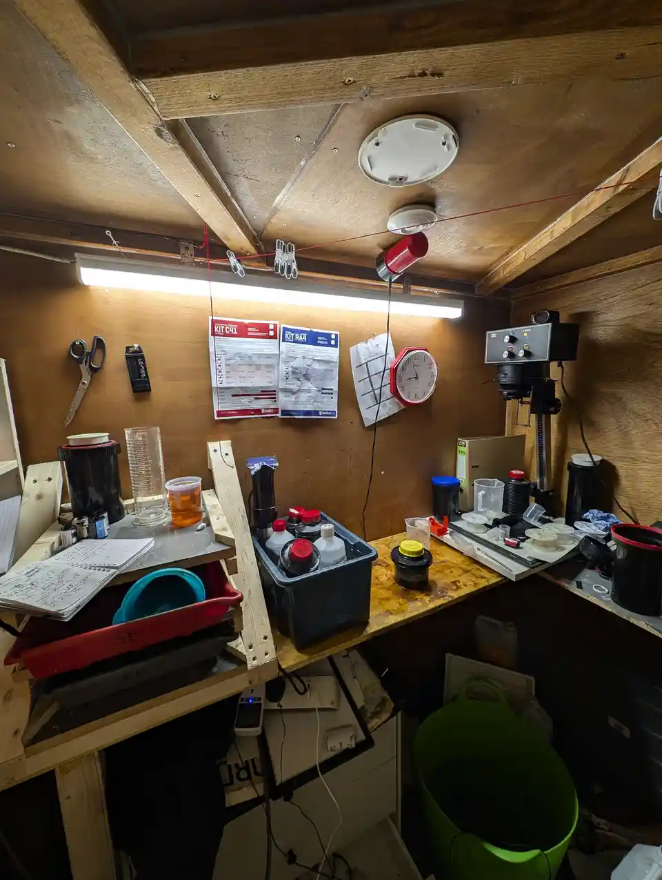 A cluttered, makeshift lab with wooden walls and ceiling, featuring a workbench covered in bottles, containers, tools, and…