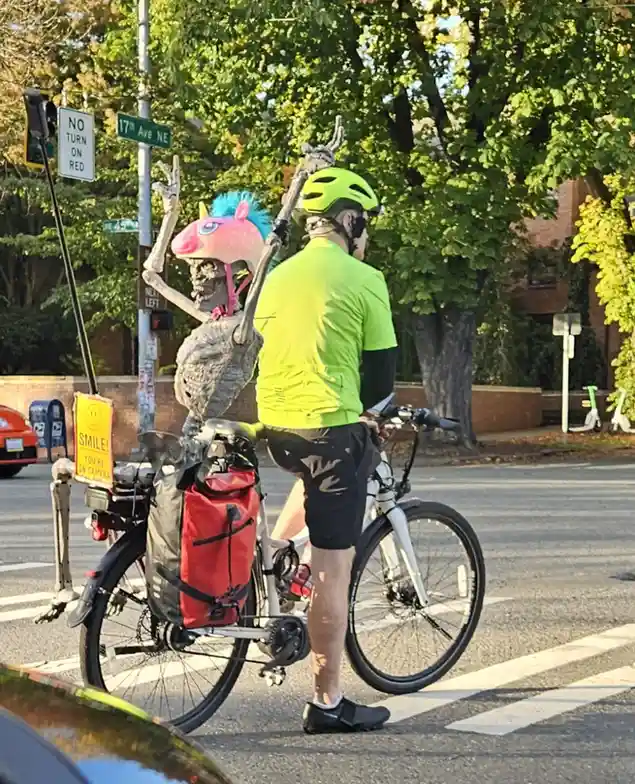A person in a bright green cycling outfit and helmet stands at a street intersection with a bicycle, facing away from the…