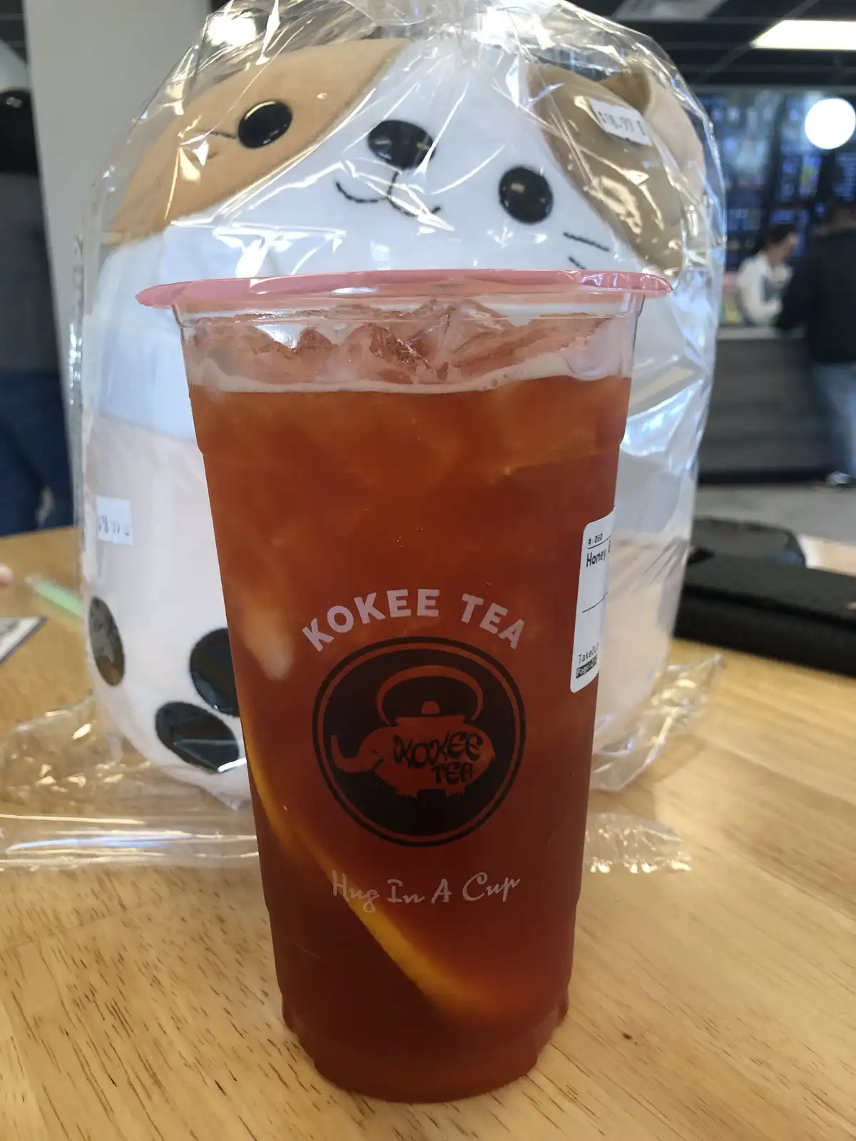 A clear plastic cup of iced tea from Kokee Tea sits on a wooden table, with a large white and tan plush dog in a plastic bag…