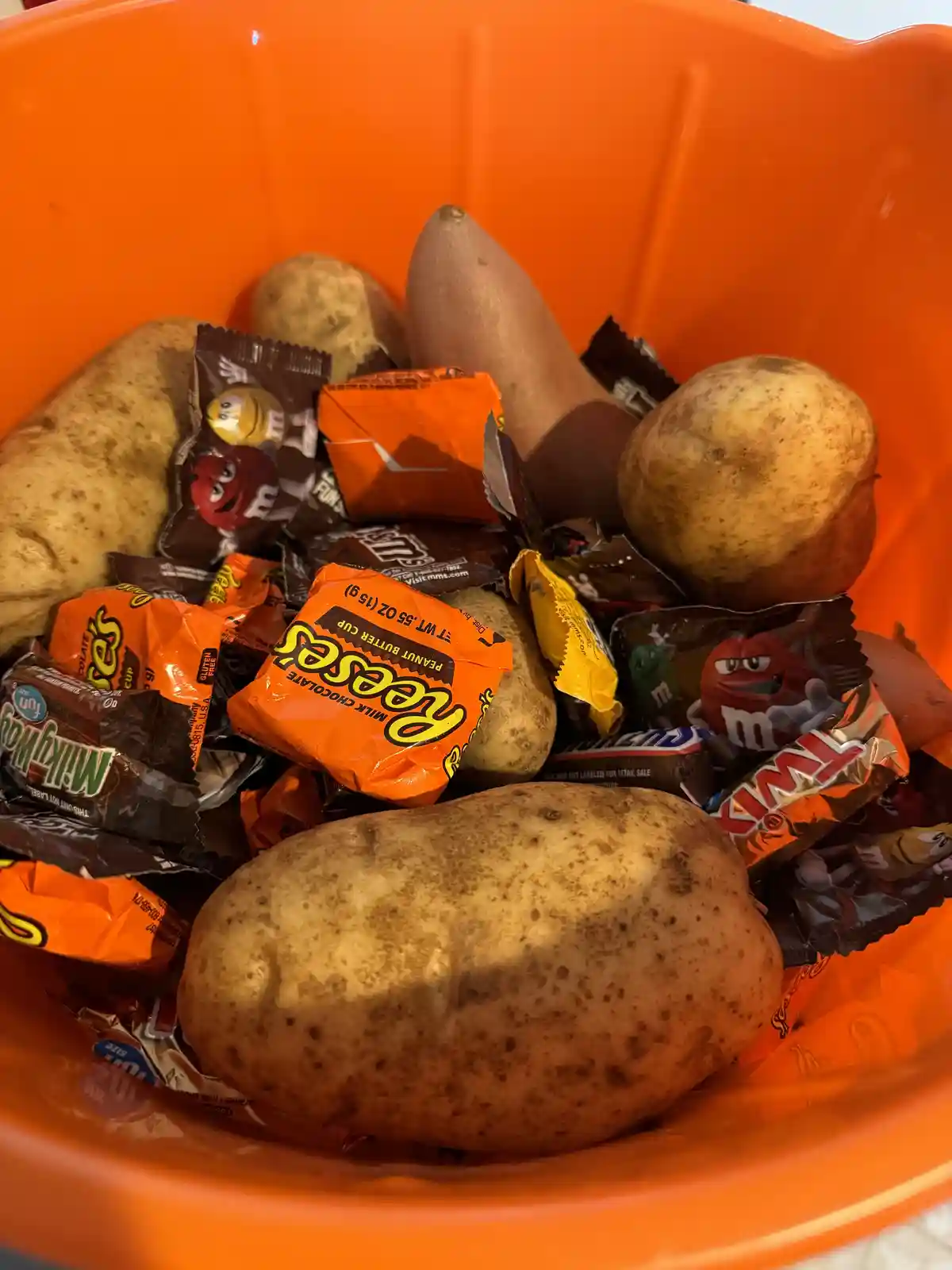 An orange plastic bowl is filled with a mix of whole potatoes and various individually wrapped candies, including Reese's…