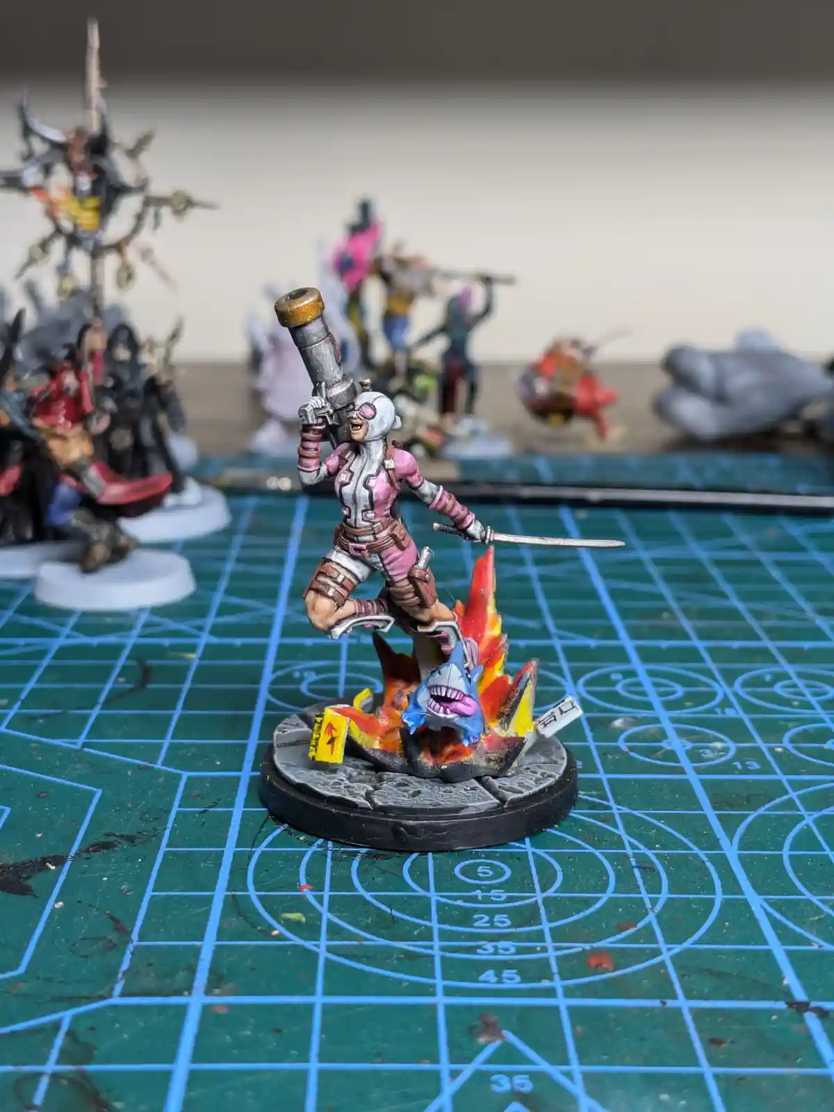 A painted miniature of a female warrior in pink and grey armor, holding a rocket launcher and a sword, stands triumphantly…