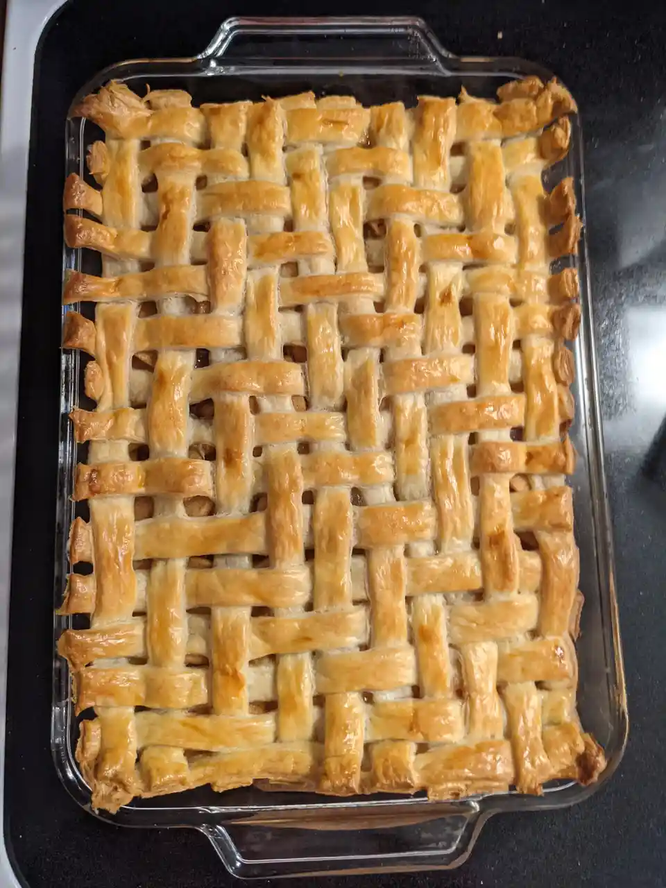 A golden-brown lattice-topped pie sits in a clear glass baking dish, its crust woven in a crisscross pattern resembling a…