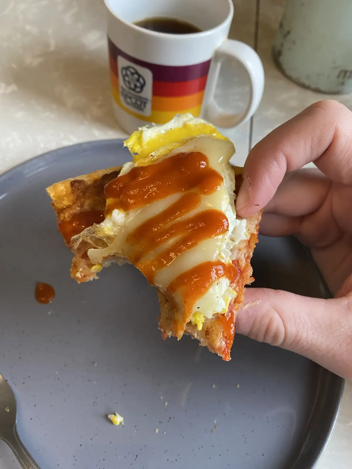 A hand holds up a partially eaten slice of breakfast pizza topped with melted cheese, a fried egg, and a drizzle of orange…
