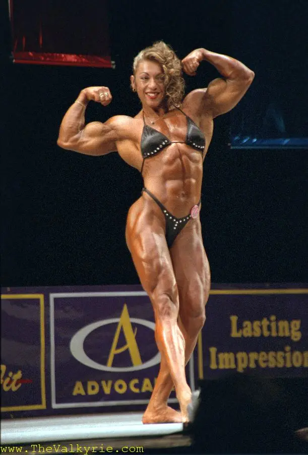 A muscular female bodybuilder in a black bikini poses on stage with both arms flexed, showcasing her defined physique under…