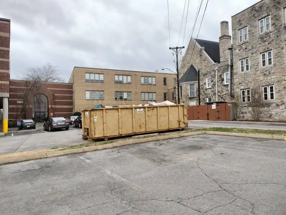 A large, rusty, tan dumpster sits in a cracked asphalt parking lot in front of brick and stone buildings under an overcast…