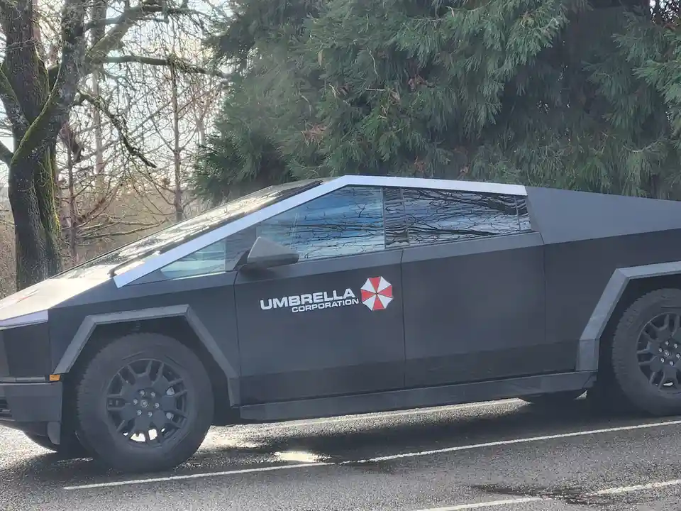 A black Tesla Cybertruck is parked on a street with trees in the background. The side of the vehicle has a white decal that…