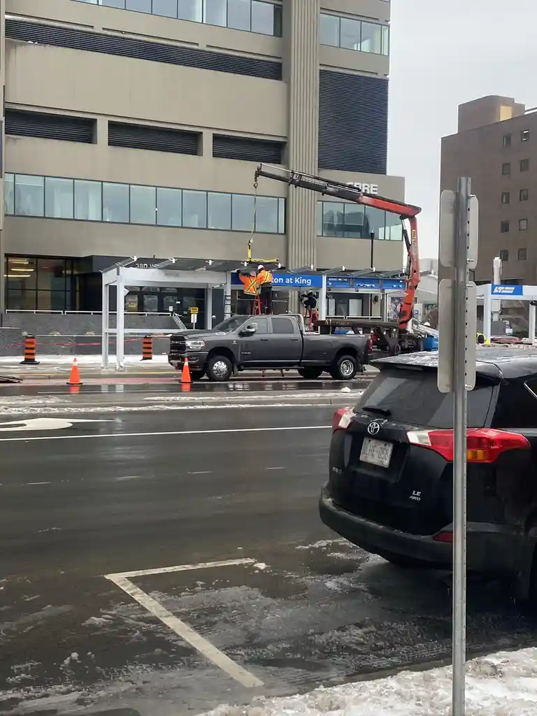 A photo shows construction workers installing a new awning over a building entrance, suspended by a crane attached to a…