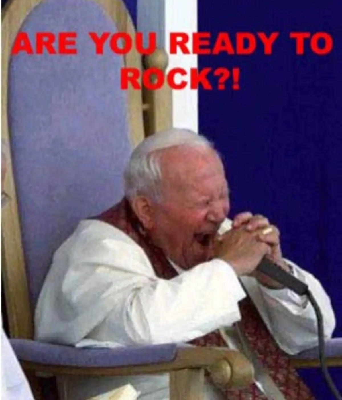 An elderly man in white clerical robes sits in a wooden chair with a purple cushion, holding a harmonica to his mouth and…