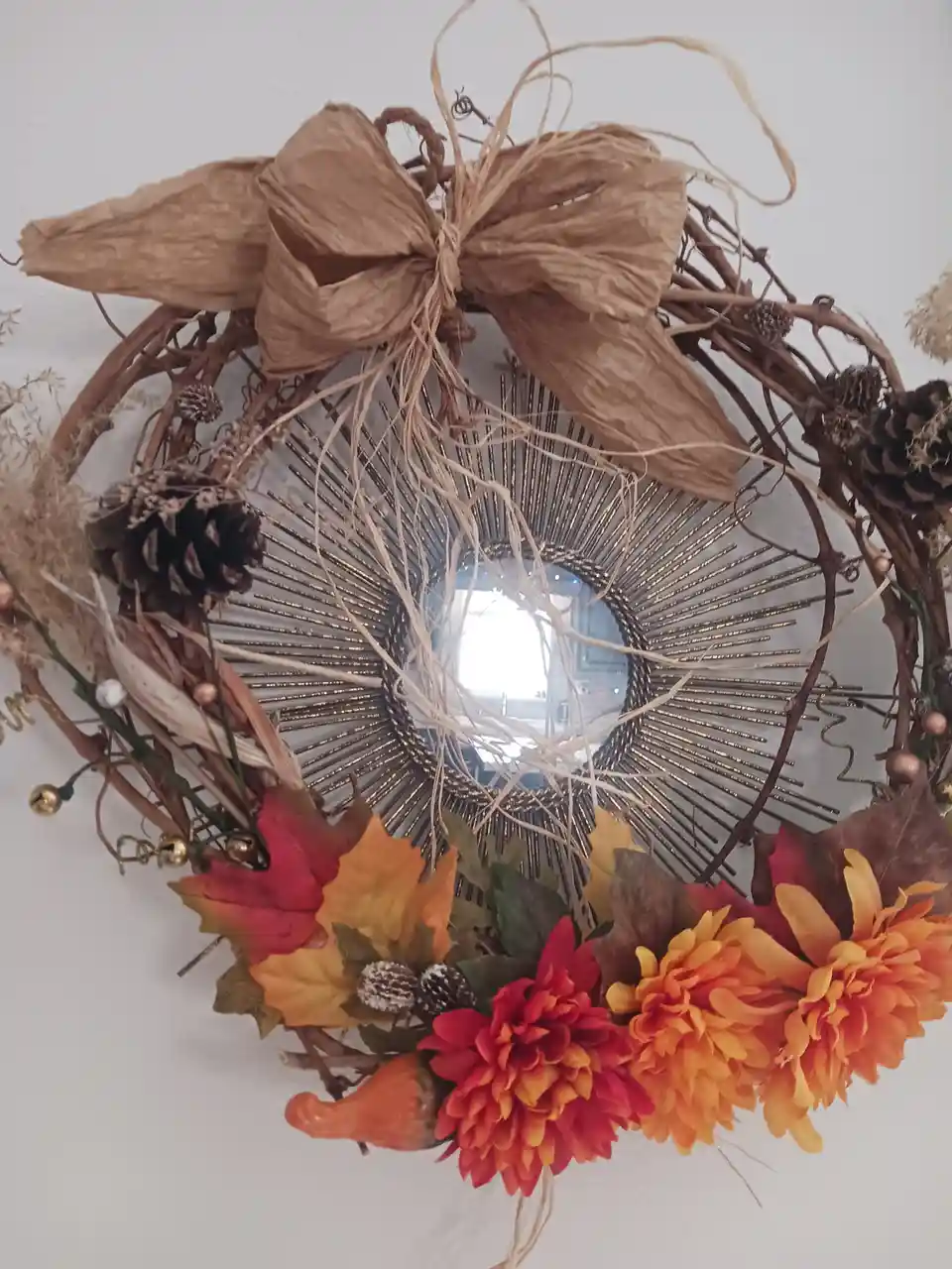 A circular decorative wreath made of brown vines, pinecones, dried leaves, orange-red artificial flowers, and a large brown…