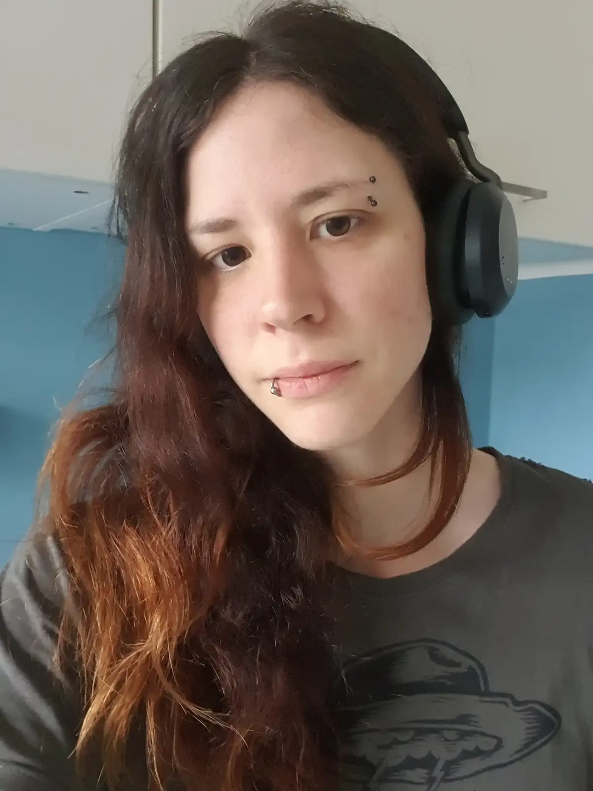 A person with long dark hair and multiple facial piercings wears large black over-ear headphones while looking directly at…