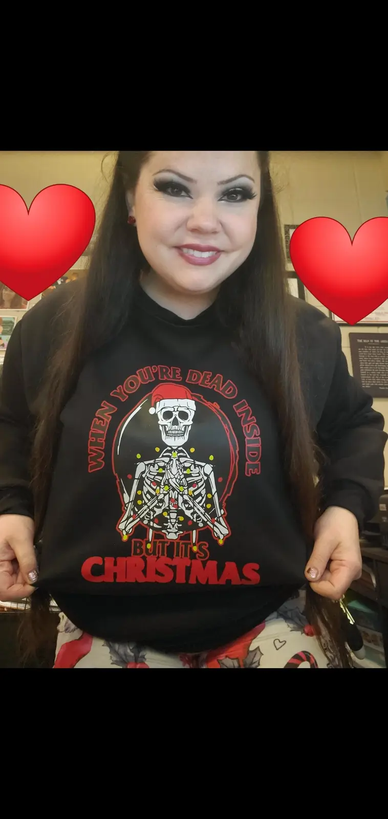 A person wearing a black sweatshirt with a graphic of a skeleton wearing a Santa hat and decorated with Christmas lights…