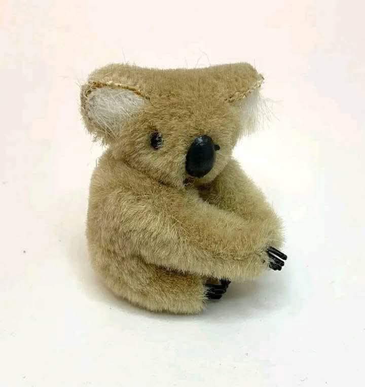 A small, handmade-looking plush koala with brown fur, black nose, and black claws sits hunched with arms crossed tightly…
