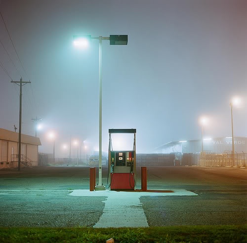 A foggy, dimly lit gas station at night with a single pump under a bright overhead light, surrounded by empty pavement and…