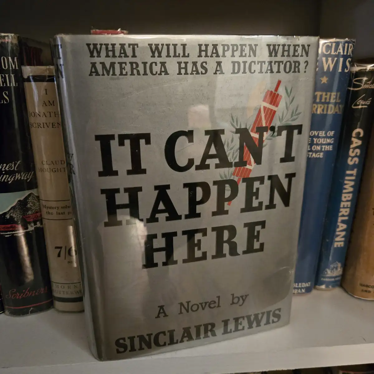A vintage book cover for Sinclair Lewis's novel 'It Can't Happen Here' is shown on a bookshelf, featuring the title and…