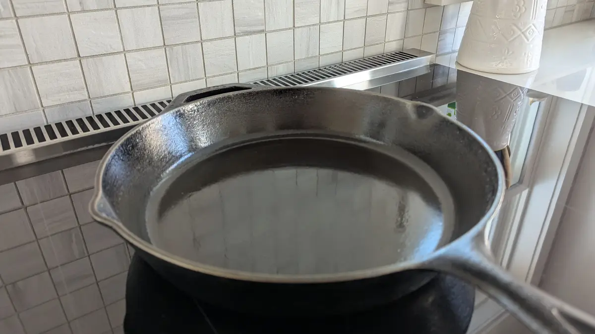 A close-up photo shows a shiny, empty cast iron skillet resting on a black stovetop in a kitchen with white tiled walls and…