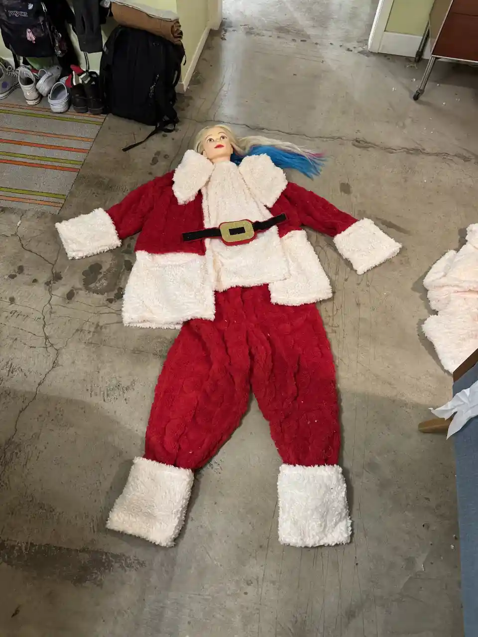 A blonde doll with blue-tipped hair lies flat on a concrete floor wearing a red and white fuzzy Santa outfit with a black…