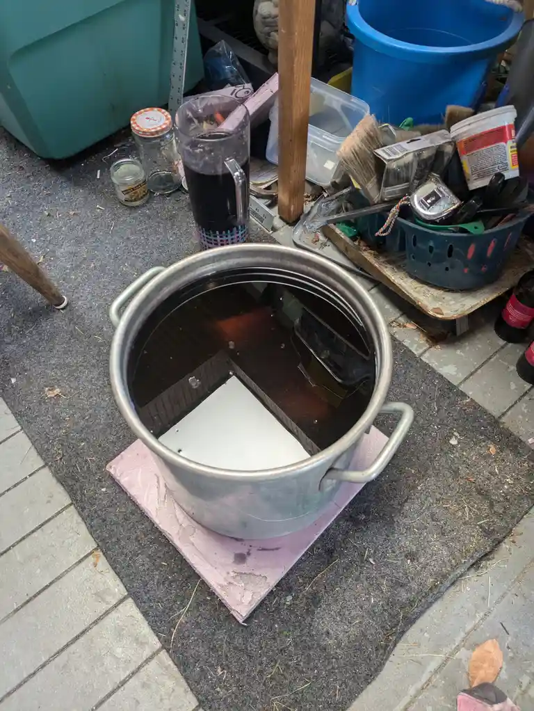A large metal pot filled with dark liquid sits on a pink mat outdoors, surrounded by clutter including a glass mug…