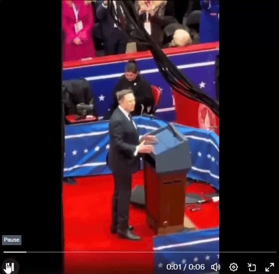 A man in a dark suit stands at a podium on a red carpeted stage with blue and white star-patterned backdrops, gesturing…